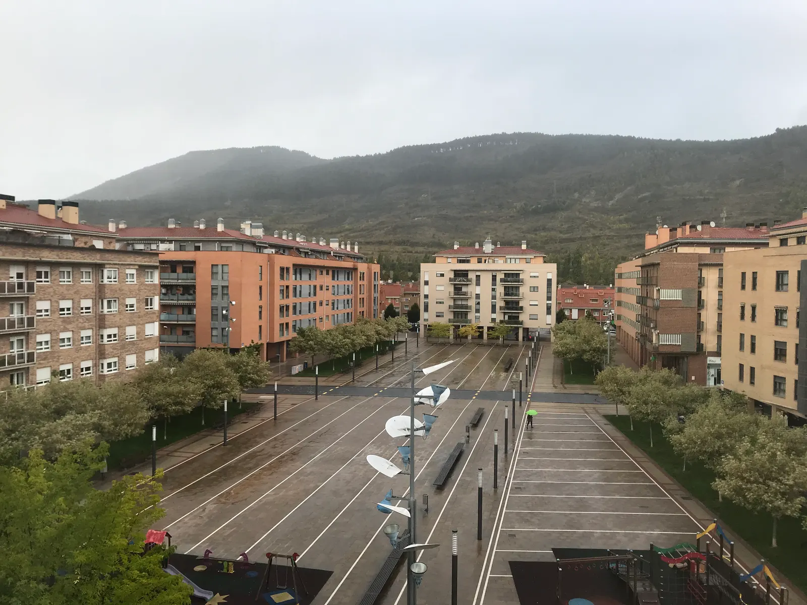 Vista de Ansoáin desde lo alto: bloques de viviendas de ladrillo a ambos lados de una plaza y el monte Ezkaba al fondo con nubes bajas