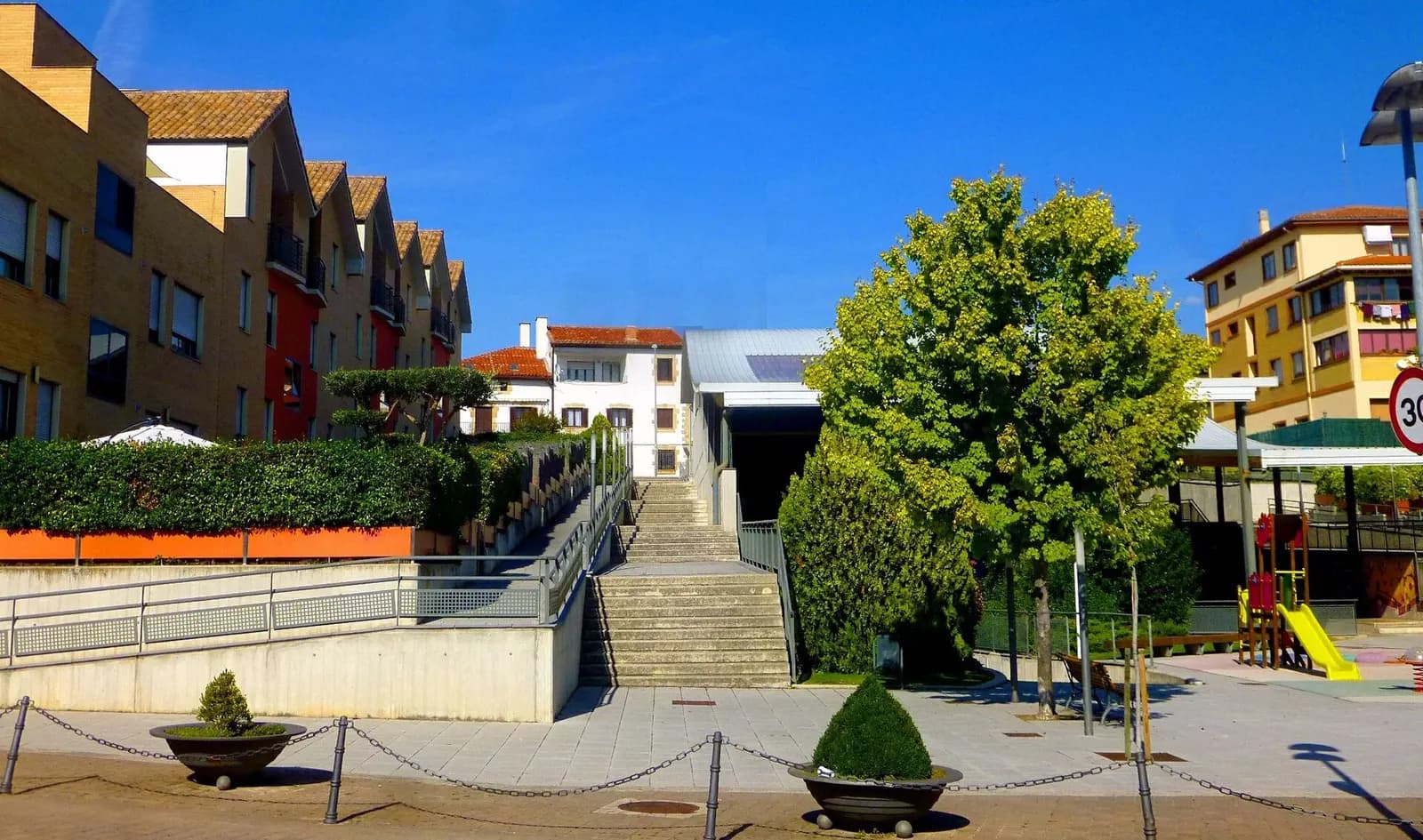 Plaza de Mutilva (Valle de Aranguren) con escaleras, jardineras, una zona de juegos infantiles y casas adosadas de ladrillo