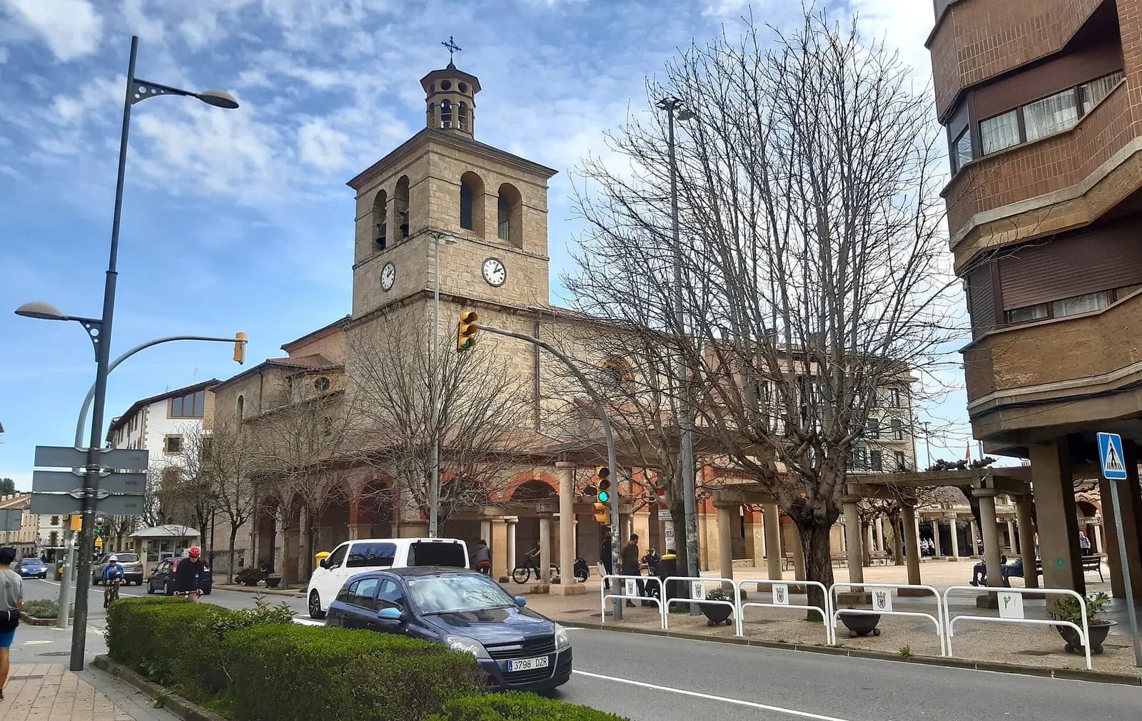 Iglesia de San Juan Evangelista de Huarte, con torre de campanario y reloj, junto a soportales y una calle con coches aparcados