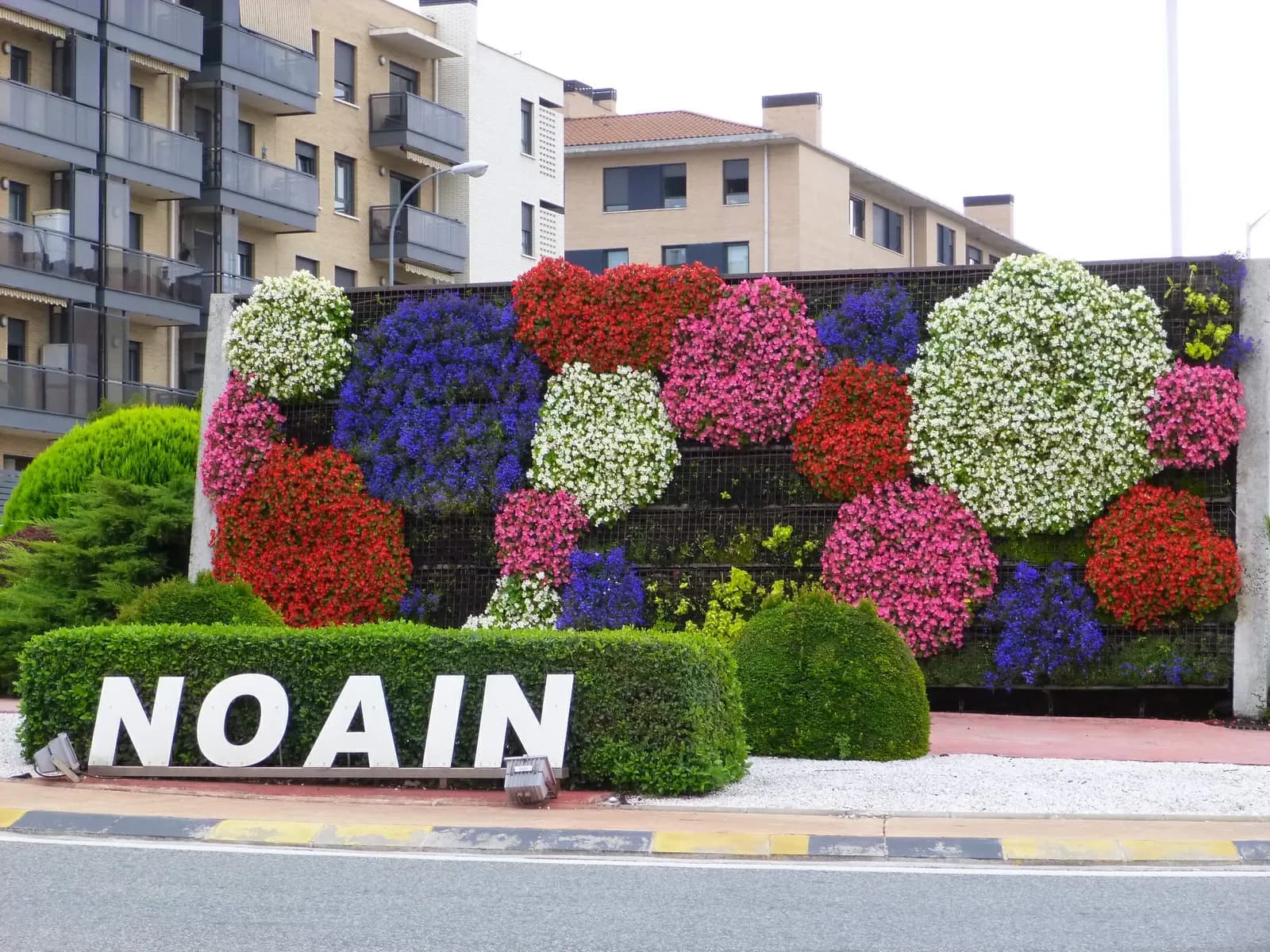 Letrero de Noáin entre setos, delante de un muro vertical de flores de colores y bloques de viviendas
