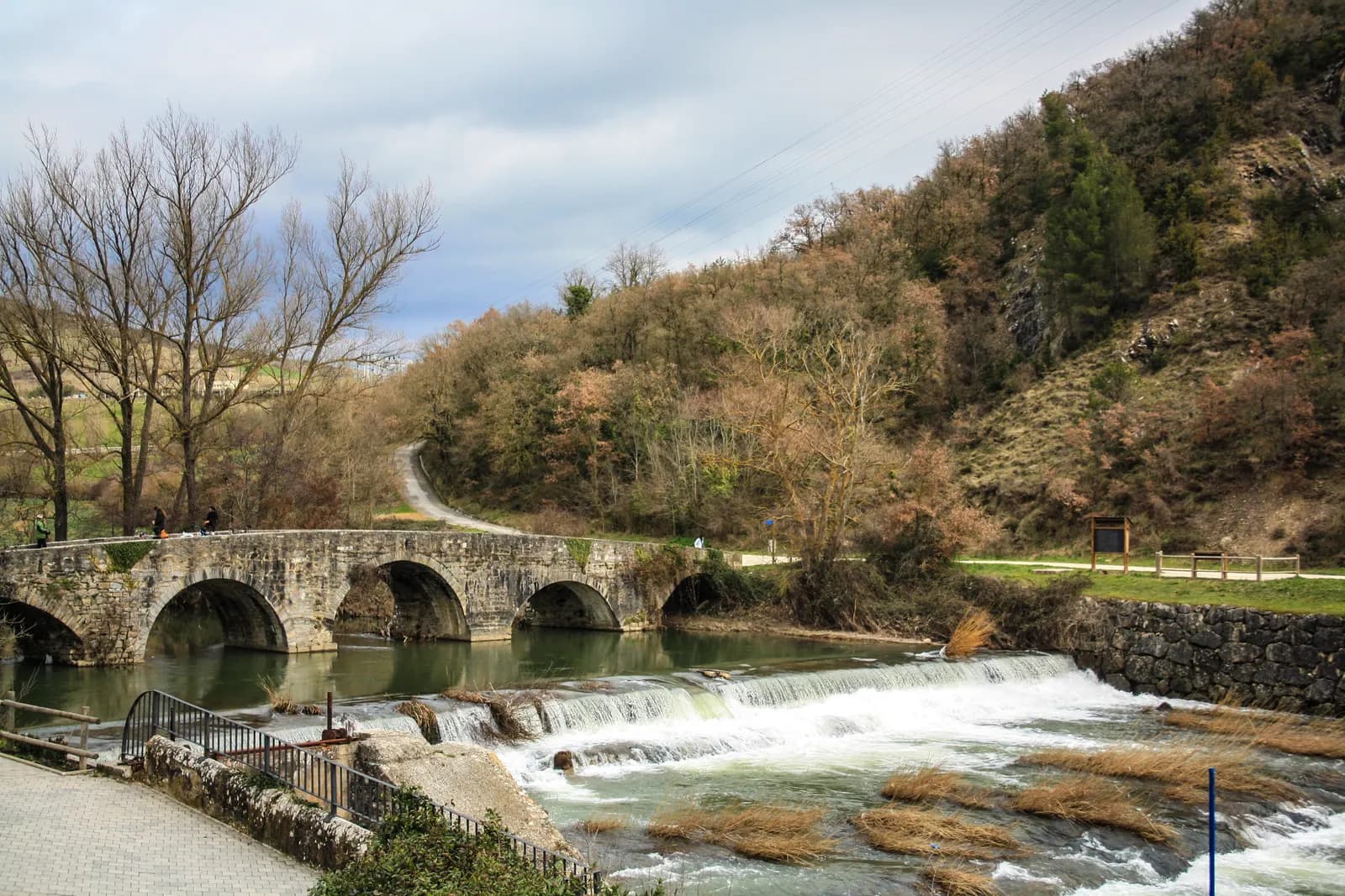 Puente medieval de piedra de la Trinidad de Arre sobre el río Ulzama en Villava, con el agua bajando por la presa y el monte al fondo