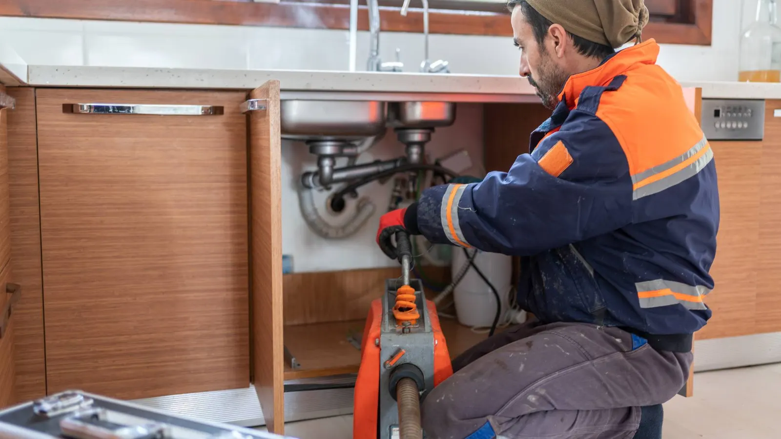 Fontanero de rodillas metiendo el cable de una máquina de muelle por el desagüe de un fregadero, con el mueble bajo abierto